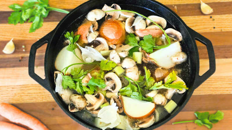 Large cast iron pot filled with beef bones, vegetables, and water, Sitting on butcher block. Carrots, parsley, and garlic on butcher block.