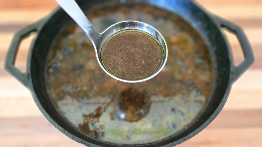 Stainless steel ladle of beef bone broth hovering over cast iron pot of broth. Sitting on top of butcher block.