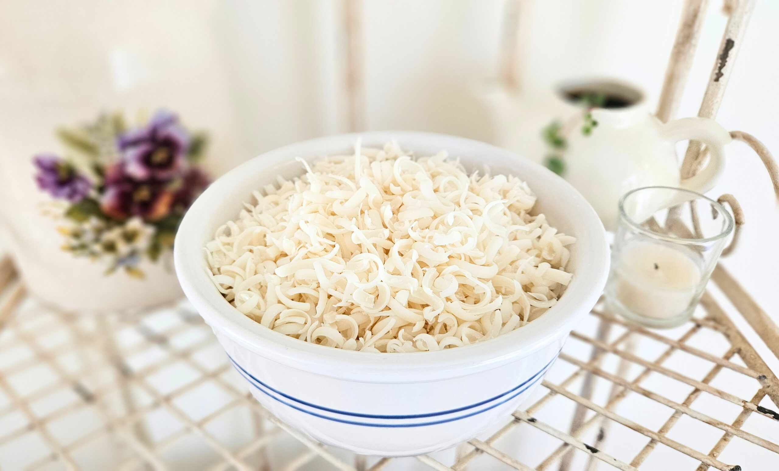 Ceramic bowl with blue striped filled with grated castile soap. Pottery and a plant in the background.