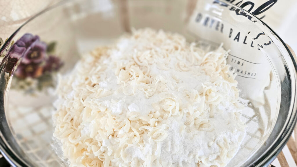 A glass bowl filled with powder laundry detergent. The detergent is a white powdery mixture with light yellow colored soap shavings.