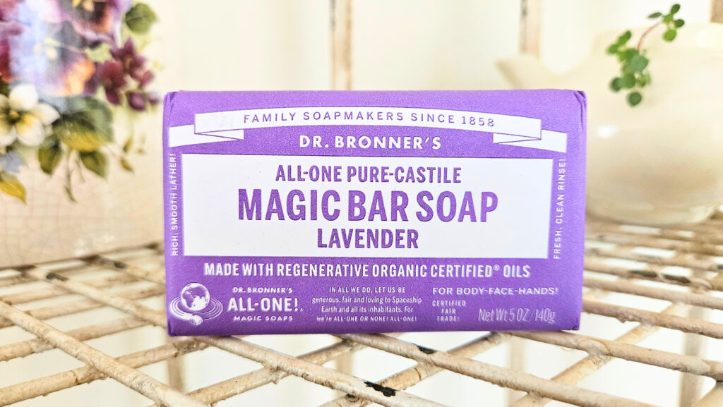 A bar of Dr. Bronner's lavender castile soap sitting on top of a metal wire shelf. Pottery and a plant are in the background.