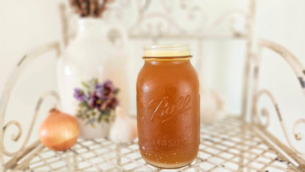 One quart size mason jar filled with bone broth. Yellow fat layer on top. The jar is sitting on top of a cream colored metal wire shelf. Cream colored pottery with purple painted flowers next to the jar of bone broth. Onions and garlic on the shelf.