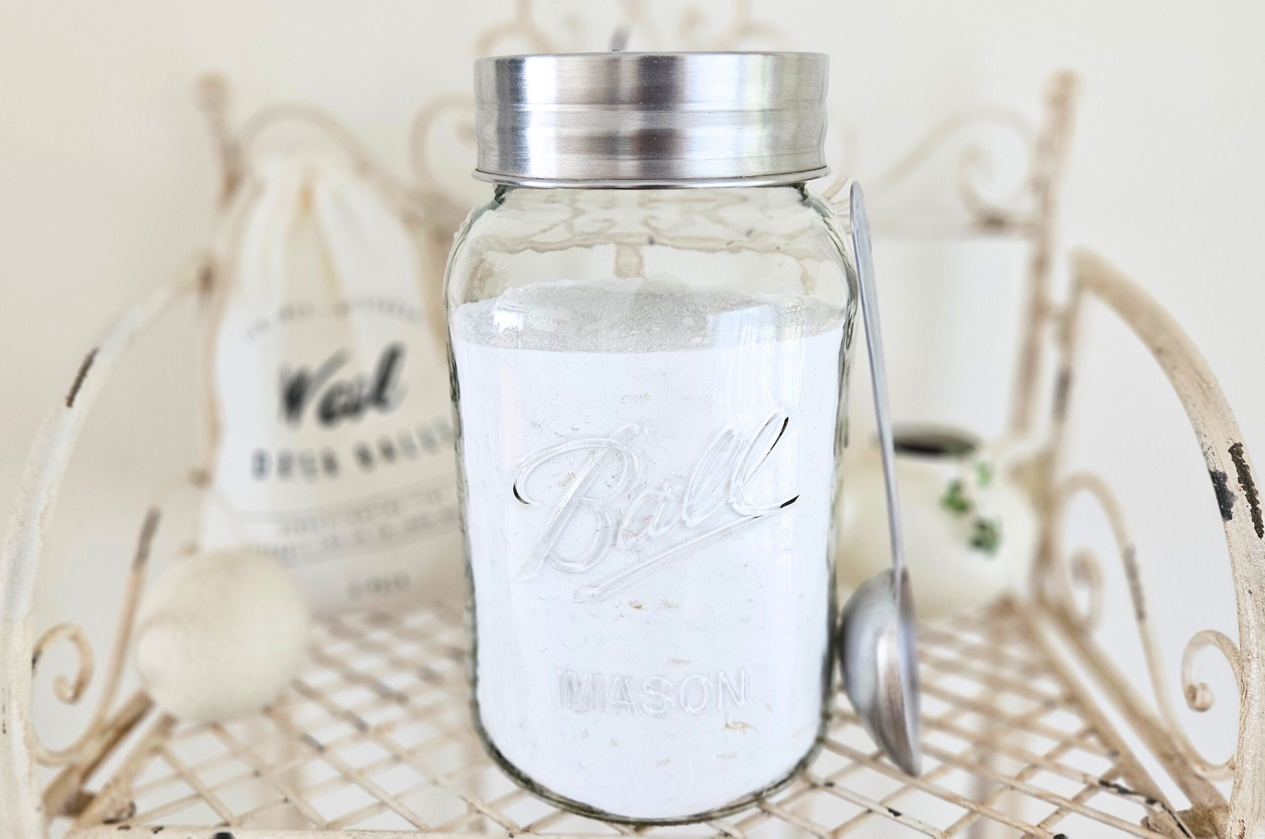 A large glass jar with a metal lid filled with powder laundry detergent. A metal scoop, wool dry ball, and plant are next to the jar.