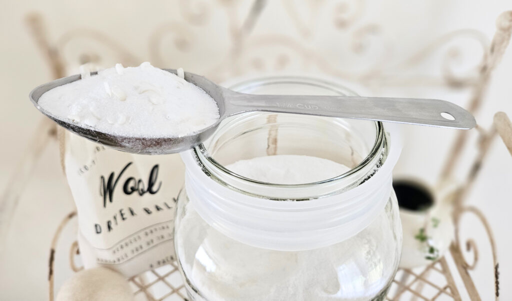 A metal scoop of powder laundry detergent. Scoop is resting on top of a large mason jar. A bag of wool dryer balls is in the background.