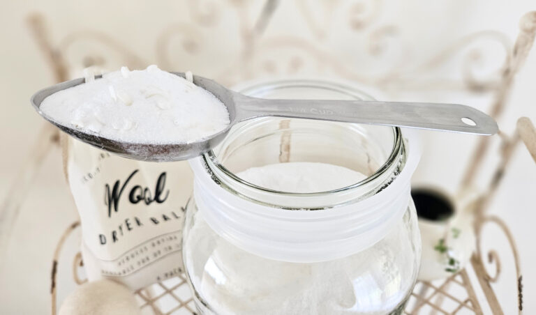 A metal scoop of powder laundry detergent. Scoop is resting on top of a gallon glass jar. A bag of wool dryer balls is in the background.