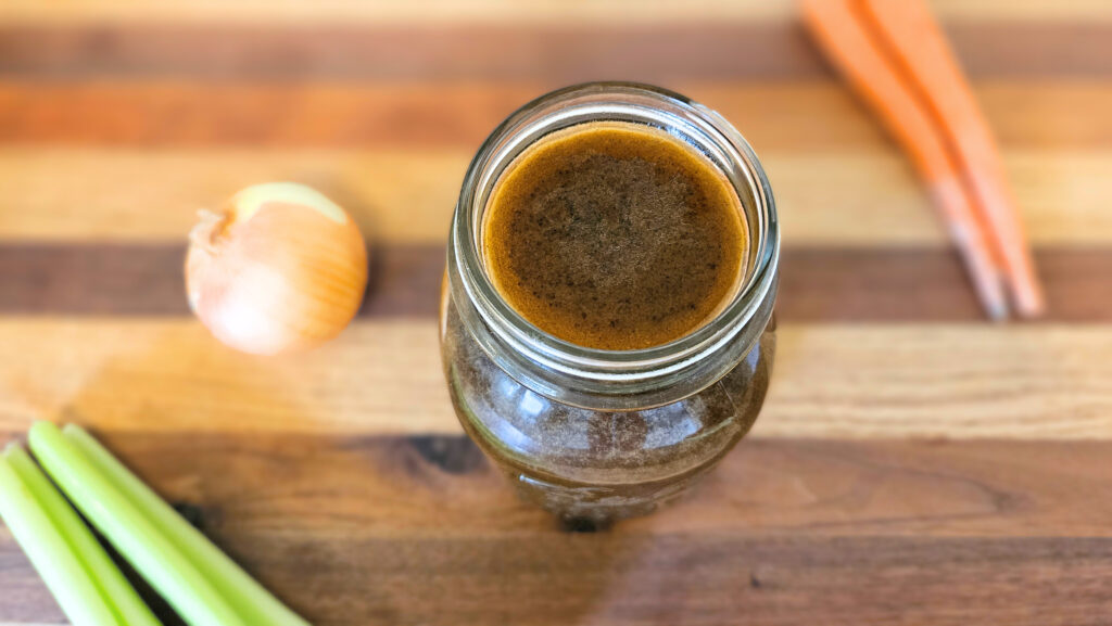 Quart size mason jar filled with beef bone broth. Sitting on top of butcher block. Carrots, celery, and an onion on the butcher block.