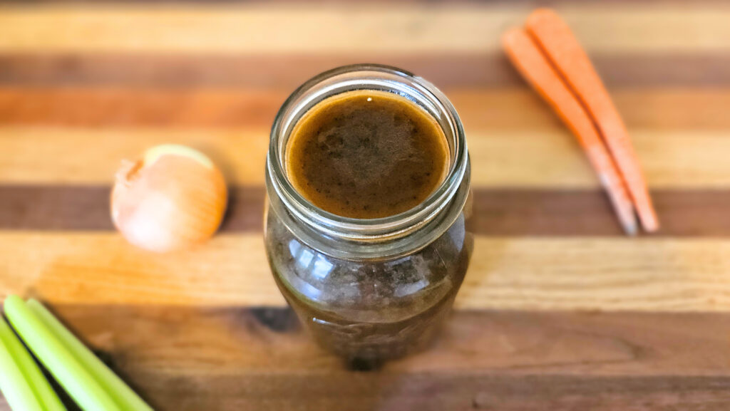 Quart size mason jar filled with beef bone broth sitting on top of butcher block. Carrots, celery, and an onion next to the jar.