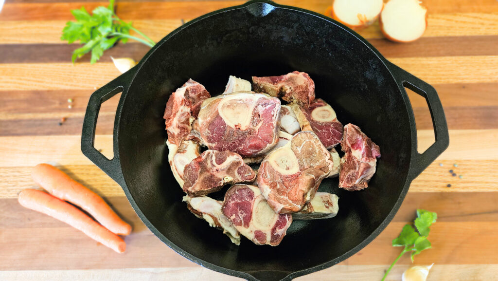 Raw beef bones in a black cast iron dutch oven. Sitting on top of butcher block with vegetables on the butcher block.