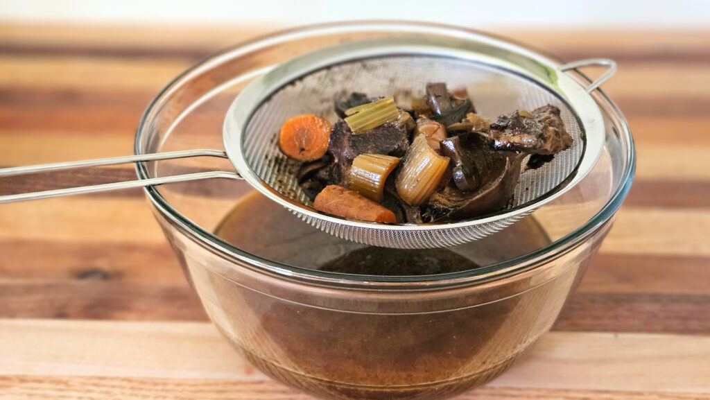 Glass bowl with stainless steel strainer on top. Beef bone broth in the both. Bones and vegetables on the strainer.