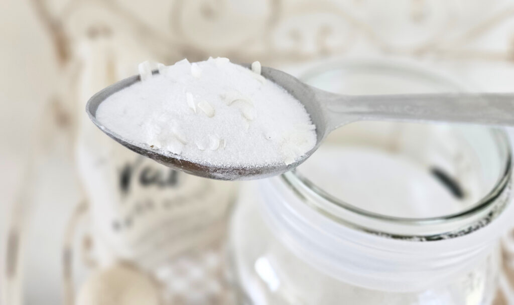 Close-up view of powder laundry detergent on a quarter cup sized metal scoop. The scoop is resting on top of a large wide mouthed glass jar.