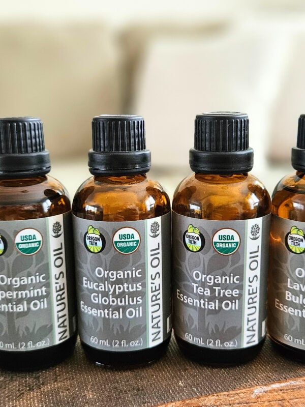 Line up of 4 essential oils: peppermint, lavender, tea tree, and eucalyptus. All sitting on a trunk with a couch and pillows in the background.