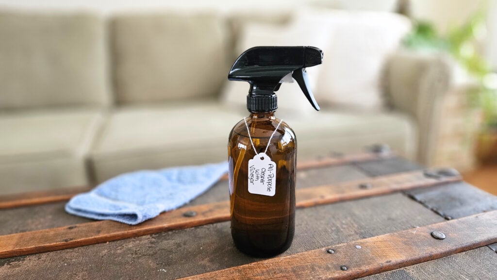 Amber glass spray bottle with all-purpose cleaner with vinegar sitting on a trunk. Blue cleaning cloth next to it.