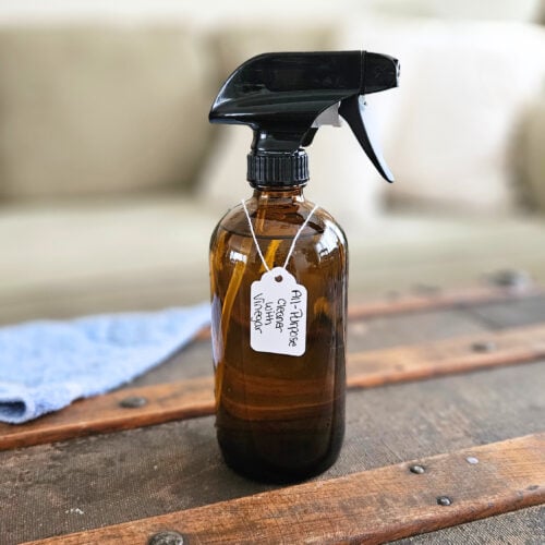 Finished product of all-purpose cleaner with vinegar sitting on top of a trunk in front of a couch with a blue cleaning cloth nearby.