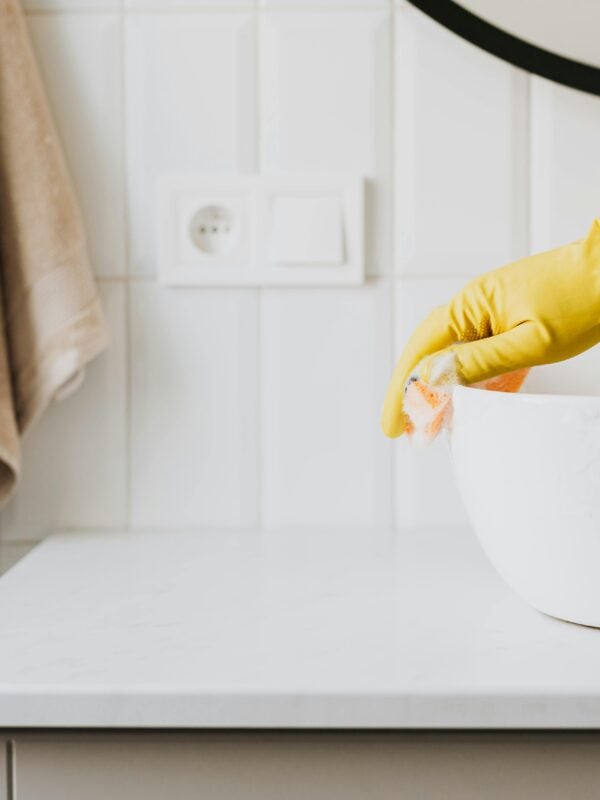 Crop anonymous person wearing yellow latex fgloves washing sink in bathroom with orange sponge and Dr. Bronner's sal suds while cleaning contemporary flat