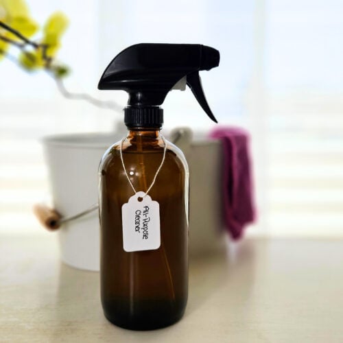 Amber glass spray bottle of DIY all-purpose cleaner. Cleaning bucket with purple cleaning cloth hanging on the side.