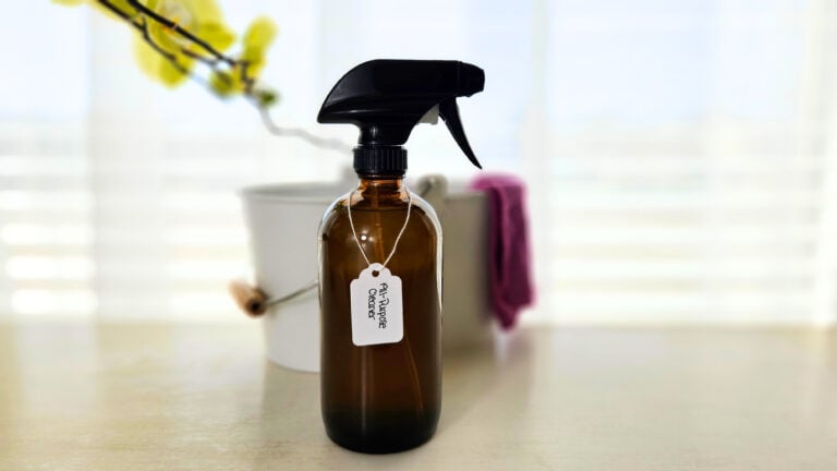 Amber glass spray bottle of DIY all-purpose cleaner. Cleaning bucket with purple cleaning cloth hanging on the side.