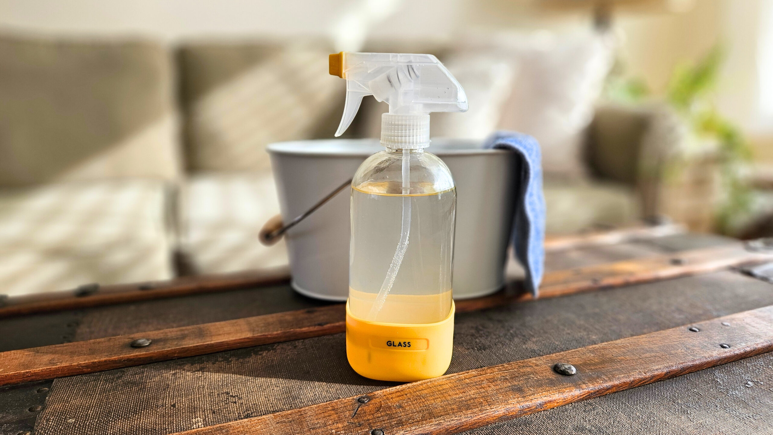 A glass spray bottle of glass and window cleaner. A white bucket with a blue cleaning cloth.