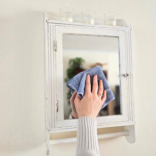 A woman's hand shown wiping the surface of a mirror with glass and window cleaner and a blue cleaning cloth.