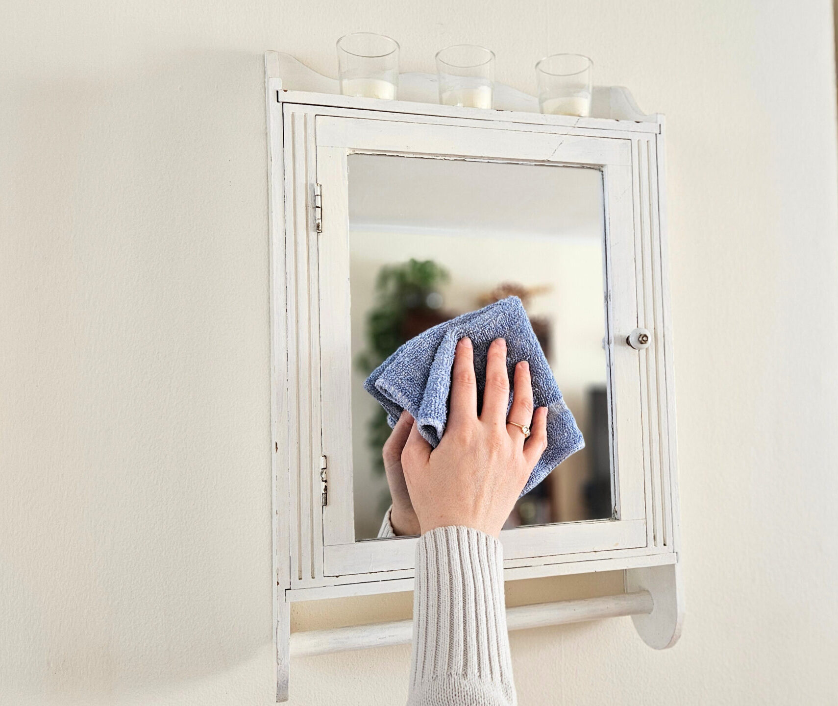 A woman's hand shown wiping the surface of a mirror with glass and window cleaner and a blue cleaning cloth.