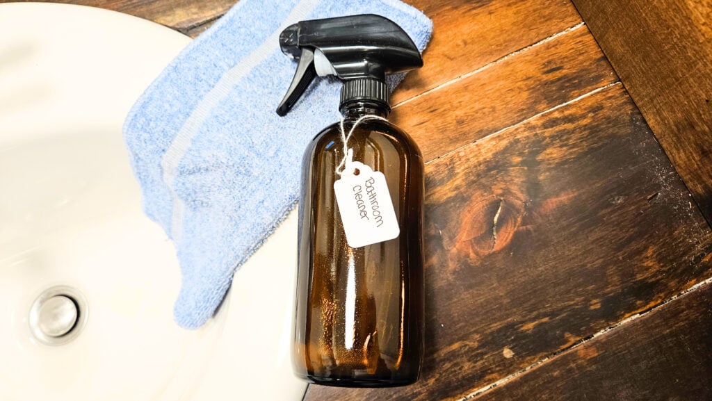 Bottle of DIY bathroom cleaner laying on a bathroom counter next to the sink with a blue cleaning cloth.