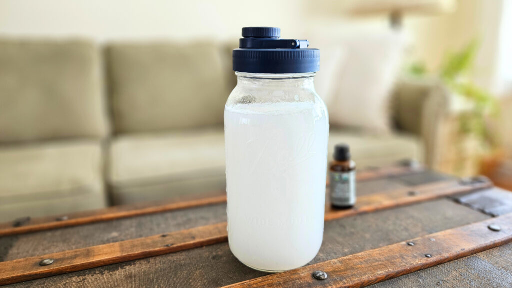 Half gallon mason jar with a blue lid filled with homemade fabric softener. Bottle of essential oil next to it.