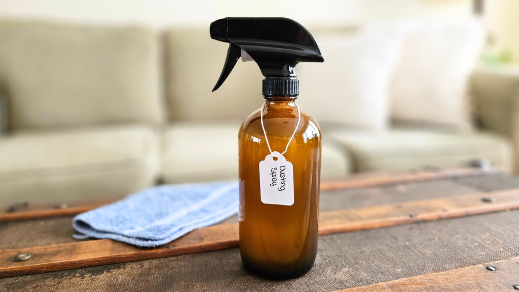 Glass bottle of dusting spray with a label and a blue cleaning cloth.