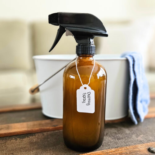 Labeled bottle of homemade dusting spray in front of a white bucket with a blue cleaning cloth.