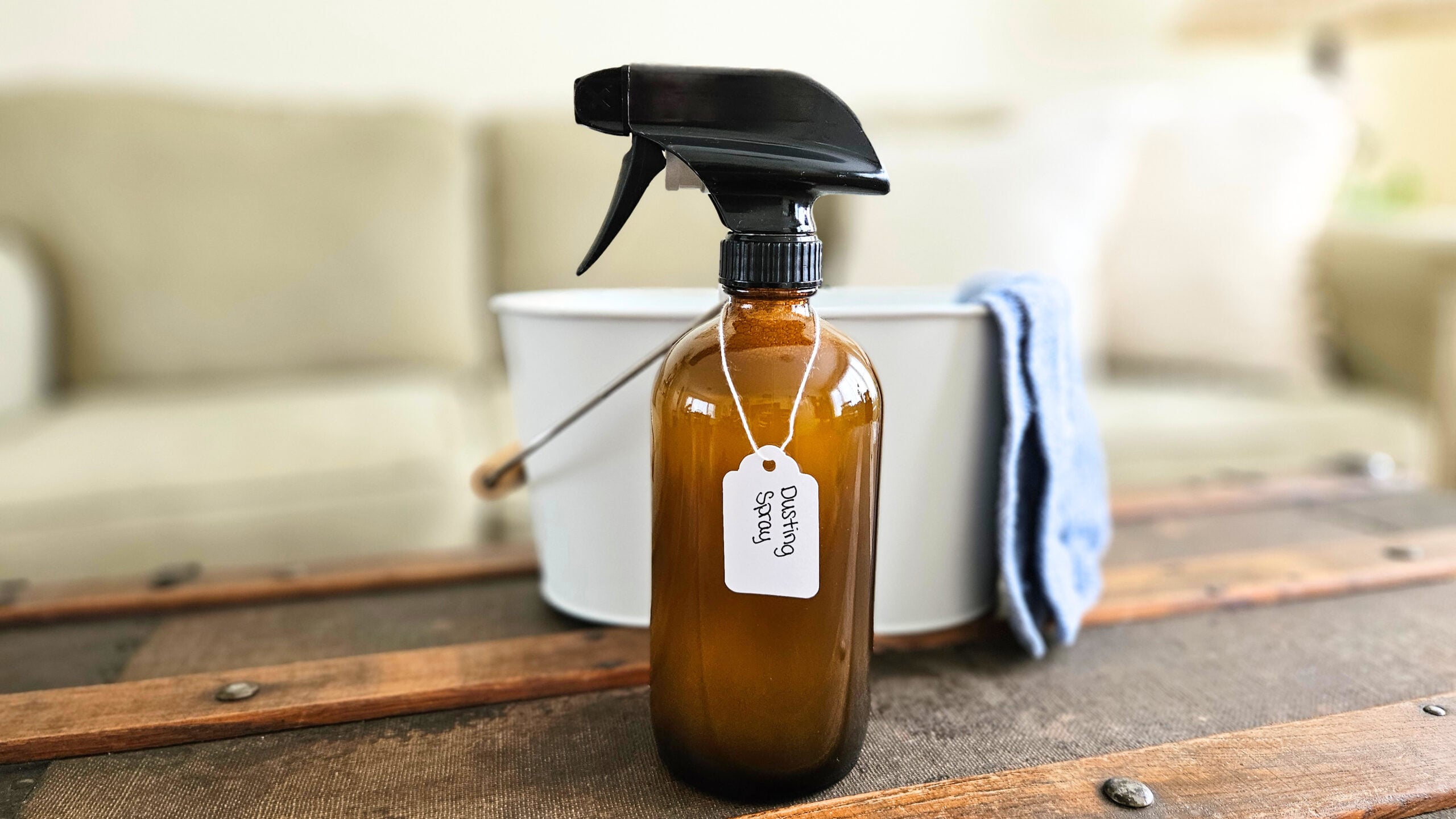 Labeled bottle of homemade dusting spray in front of a white bucket with a blue cleaning cloth.