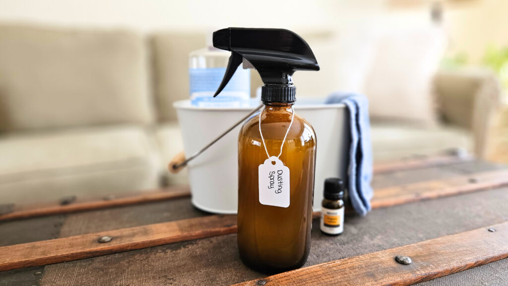 Glass bottle of homemade dusting spray with castile soap in a white bucket and a bottle of lemon essential oil.