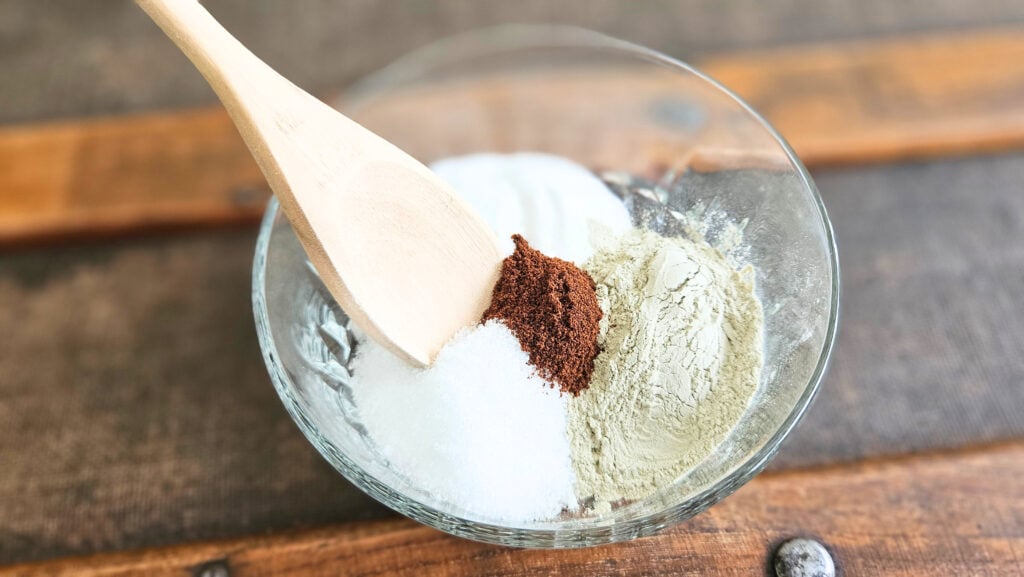 Glass bowl filled with dry ingredients for homemade tooth brushing powder and a wooden spoon.